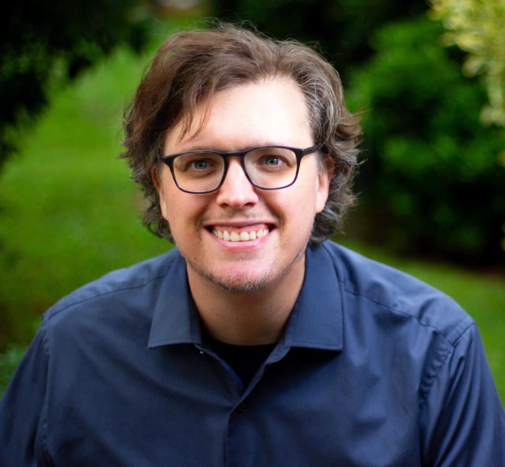 A person with curly hair and glasses smiling at the camera, wearing a navy blue shirt, with a green blurred background of foliage.