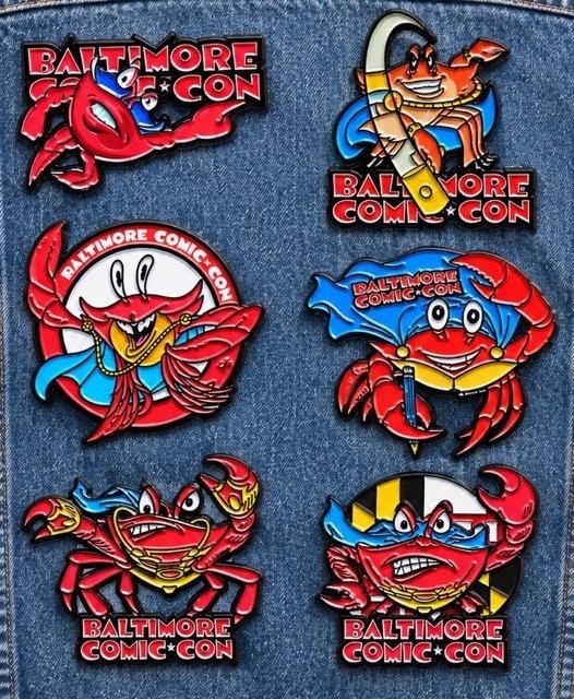A collection of colorful enamel pins featuring cartoon-style crabs and the Baltimore Comic-Con logo, arranged on a denim background. The designs showcase different styles and expressions of the crabs, some holding objects like a pencil or wearing accessories.