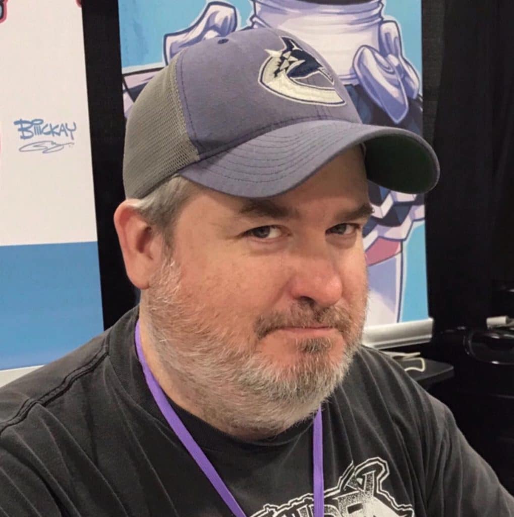 Man with a gray baseball cap and a beard, looking at the camera with a serious expression, seated in front of a colorful background at a convention.
