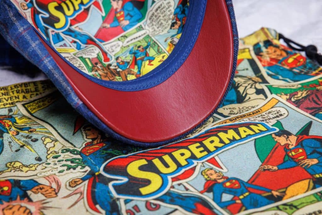 Close-up of a Superman-themed cap with a red brim and comic book design inside, resting on a fabric with colorful Superman comic panels.