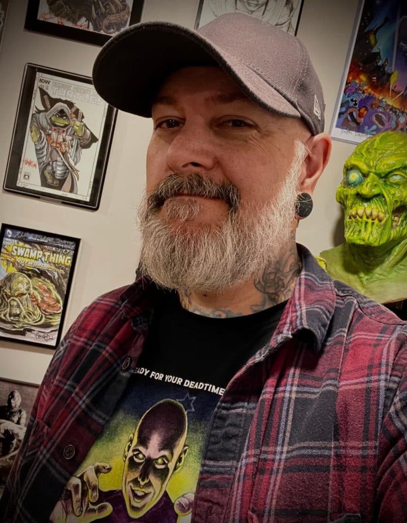 A man with a beard and a cap is smiling in a room decorated with comic book art and collectibles, including a green monster figure.