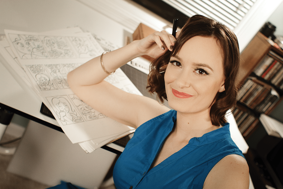 A woman with wavy hair wearing a blue top smiles while holding a pen beside a work desk covered with comic sketches.