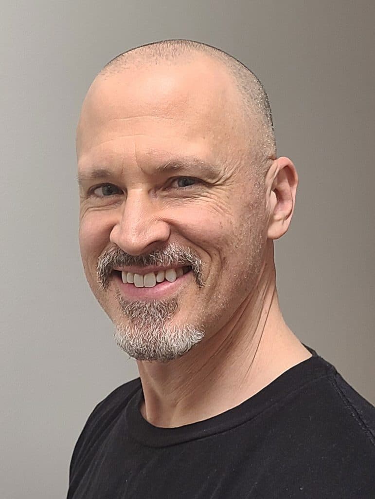 A smiling man with a shaved head and a greying goatee, wearing a black t-shirt, against a light background.