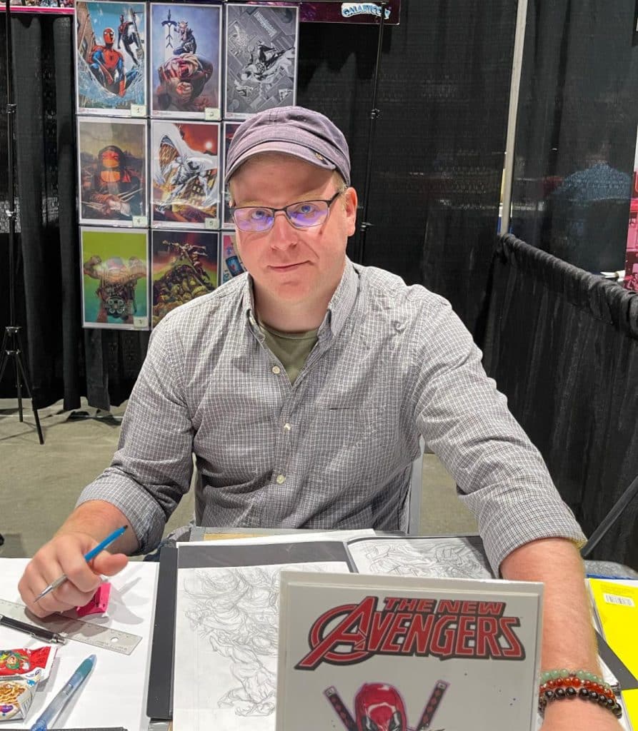 A seated artist wearing glasses and a cap, working at a table with sketches and supplies, with a display of comic art behind him.