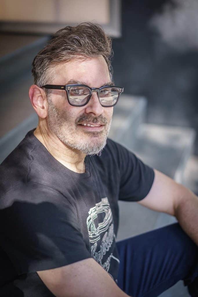 A middle-aged man with glasses and a beard, sitting on stairs outdoors, smiling at the camera, wearing a black t-shirt.