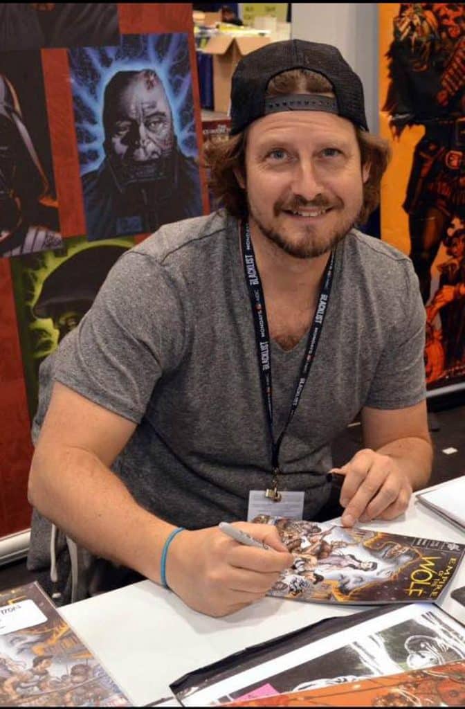 A man in a gray t-shirt and a baseball cap is smiling and signing a comic book at a convention table, with colorful artwork visible in the background.