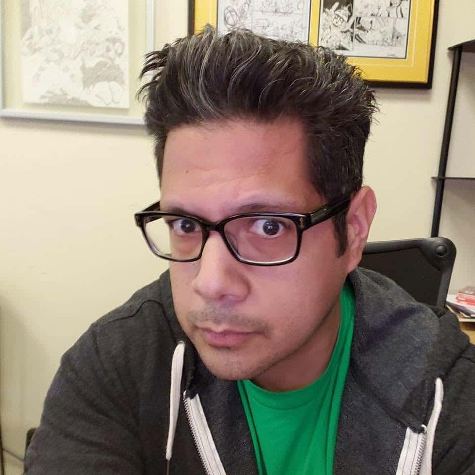 A close-up portrait of a man with dark, spiked hair and glasses, wearing a gray hoodie over a green shirt. He is giving a serious expression, with a blurred office background featuring framed comic art.