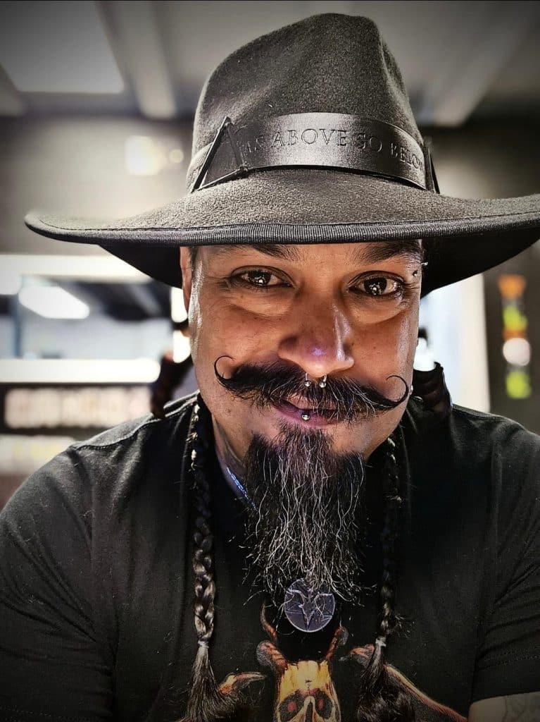 A man wearing a wide-brimmed black hat and a graphic t-shirt, with braided hair, a distinctive mustache, and multiple facial piercings, smiling at the camera.