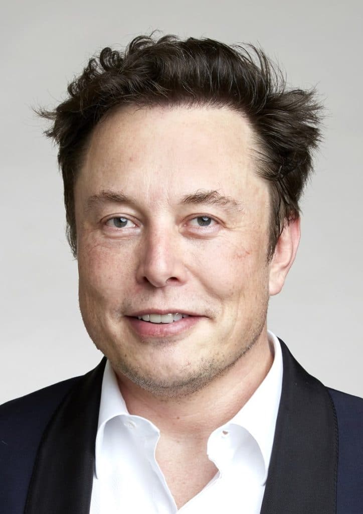 Close-up portrait of a man with tousled dark hair and light blue eyes, wearing a black suit jacket over a white shirt, smiling softly against a neutral background.
