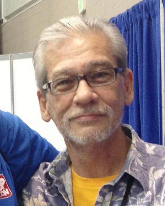 A close-up portrait of an older man with gray hair and glasses, smiling, wearing a floral shirt and a yellow t-shirt underneath, set against a blue curtain backdrop.