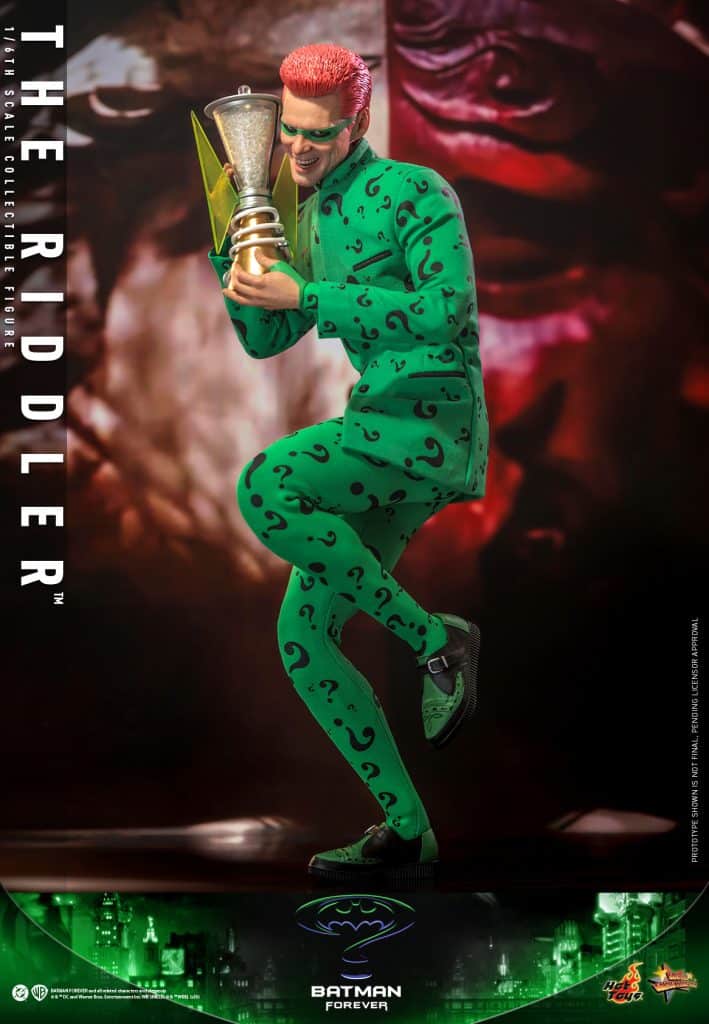 A collectible figure of The Riddler from Batman Forever, wearing a green suit with question mark patterns, holding a trophy and smiling, set against a dramatic background.