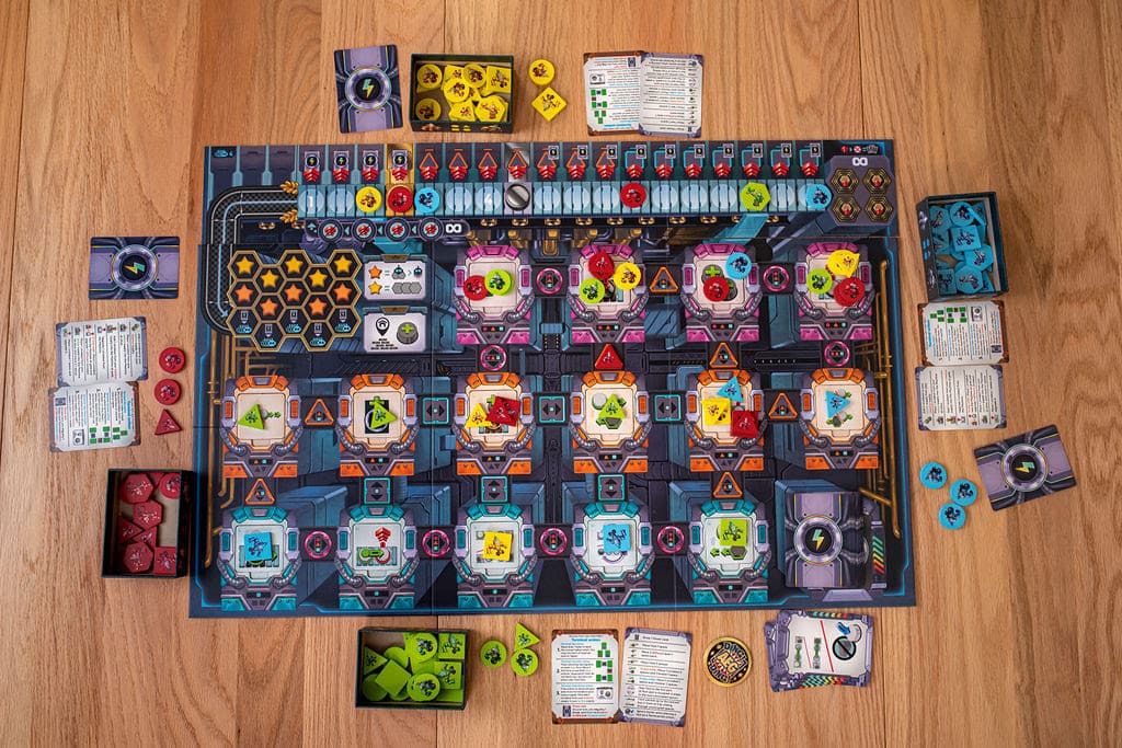 A tabletop game board set on a wooden surface, featuring a colorful layout with various tokens, cards, and dice scattered around it. The board includes sections marked with icons and hazard symbols, suggesting a strategic gameplay involving resource management or problem-solving.