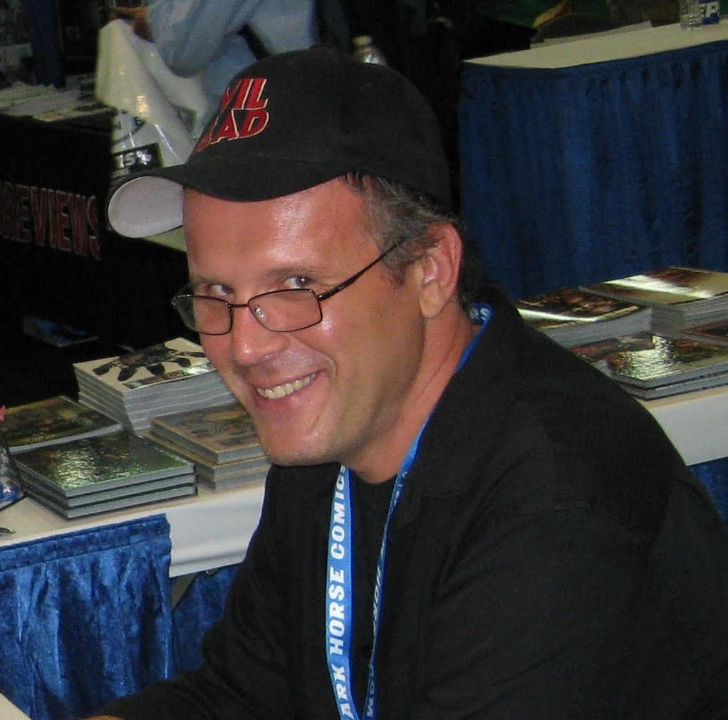 Smiling man wearing glasses and a cap with 'Evil Dead' logo, sitting at a table with comic books in the background.
