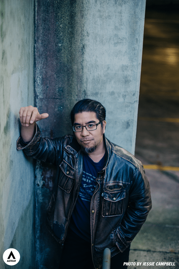 A man with glasses and long hair stands against a textured concrete wall, wearing a leather jacket and a dark shirt, giving a thumbs-up with one hand.