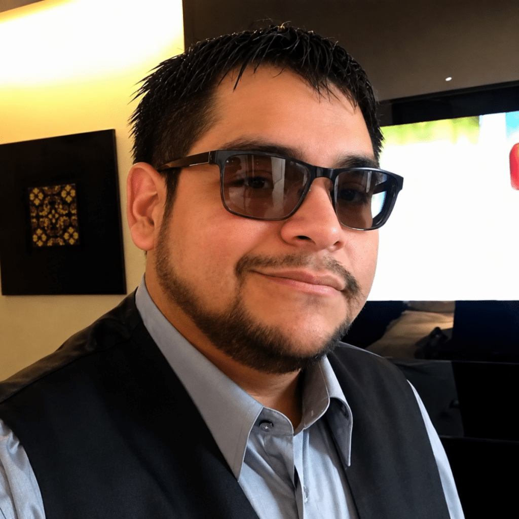 A man wearing sunglasses and a vest smiles for a selfie in a modern interior setting.