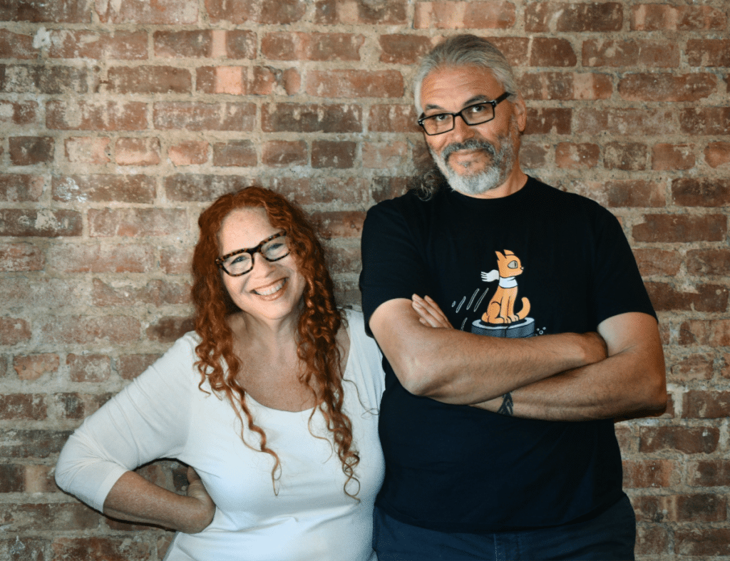 Two people standing together in front of a brick wall, one with long curly red hair and glasses, the other with longer gray hair and glasses. The woman is smiling and wearing a white top, while the man is wearing a black t-shirt with an illustrated cat design.
