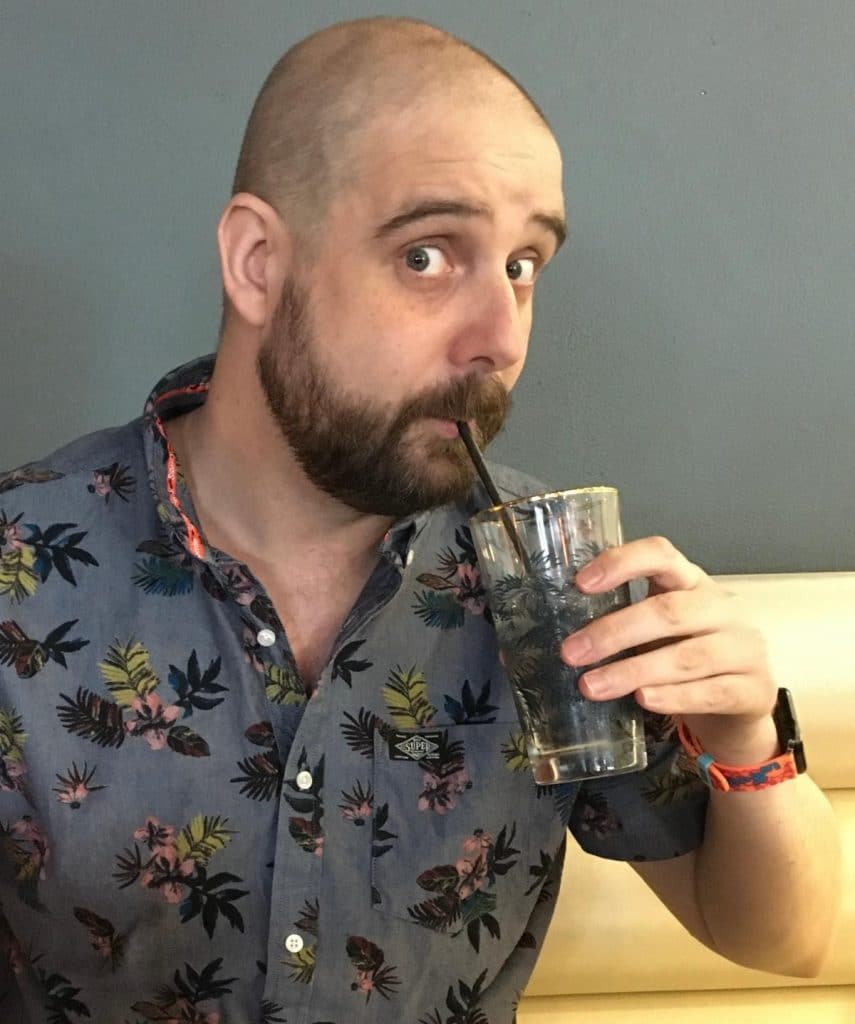 A man with a shaved head and a beard is sipping a drink through a straw. He is wearing a colorful floral shirt and has a playful expression.