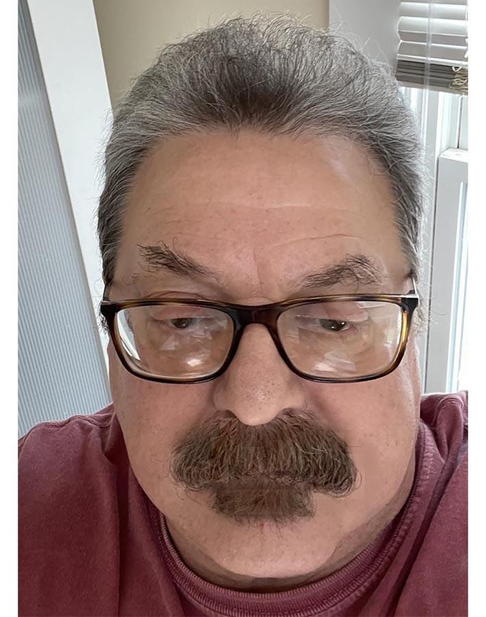 A close-up portrait of a man with gray hair and a mustache, wearing glasses and a burgundy shirt.
