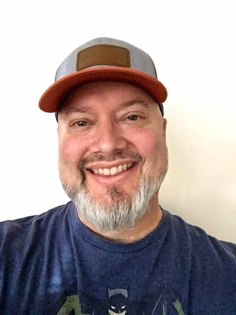 A smiling man with a gray beard wearing a cap and a blue t-shirt with a graphic design.