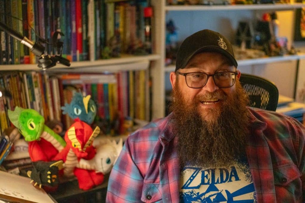 A smiling man with a thick beard wearing a blue Zelda t-shirt and plaid shirt sits in front of a shelf filled with books and collectibles. Plush dragon figures are positioned next to him.