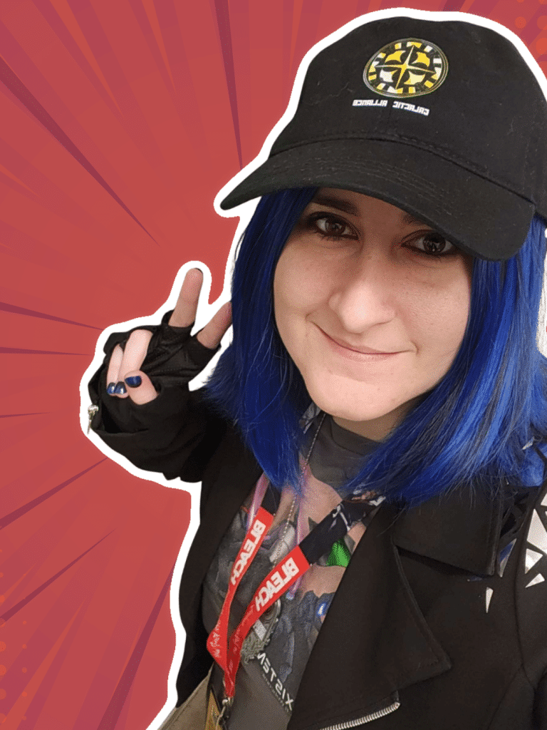 A person with blue hair wearing a black cap and a stylish black jacket, smiling and making a peace sign. They have a lanyard with badges around their neck.