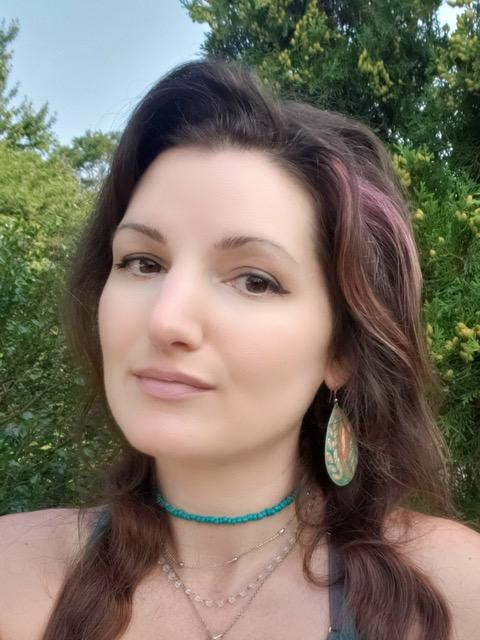 A woman with long, wavy brown hair, wearing earrings and layered necklaces, poses outdoors with greenery in the background.