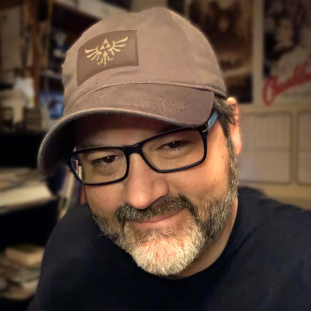 A smiling person wearing a brown cap featuring a logo from a popular video game series, with glasses and a short beard, in front of a cluttered background.