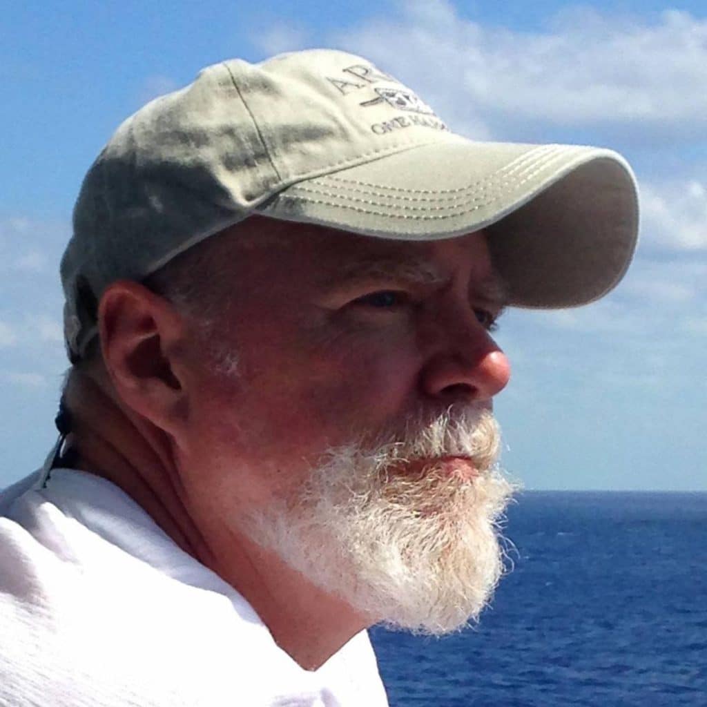 Profile of a man with a beard wearing a beige cap, looking thoughtfully towards the ocean with a blue sky in the background.