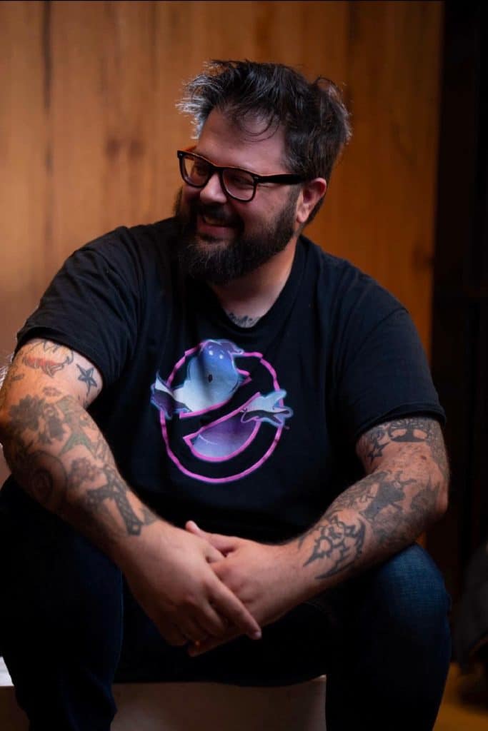 A smiling man with a beard and glasses sitting on a stool, wearing a black t-shirt featuring a colorful ghost design. He has numerous tattoos visible on his arms and is positioned in a warmly lit indoor setting.