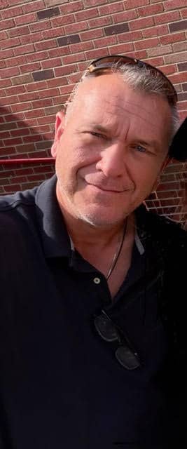 A man smiling in front of a brick wall, wearing a black polo shirt and sunglasses on his head.