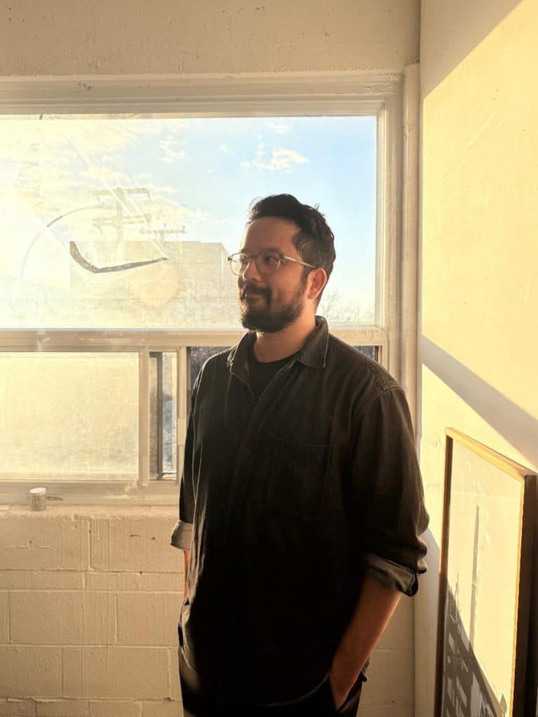 A man standing by a window with sunlight streaming in, wearing glasses and a dark shirt, with a thoughtful expression.