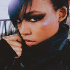 Close-up portrait of a person with dramatic eye makeup, wearing a black high-collared jacket.