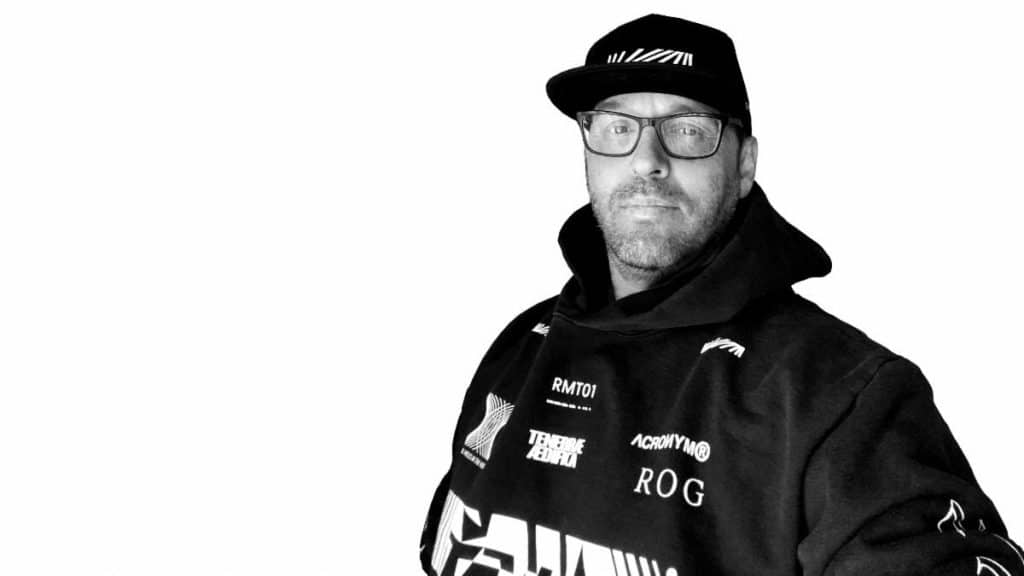 A man wearing glasses and a black hoodie with various logos, posing against a plain white background.