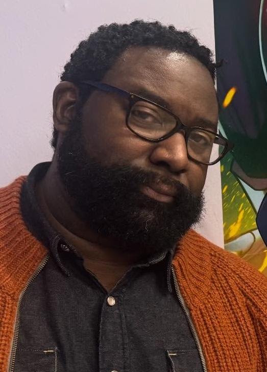 A man with glasses and a large beard, wearing an orange sweater over a dark shirt, poses thoughtfully in front of a colorful background.