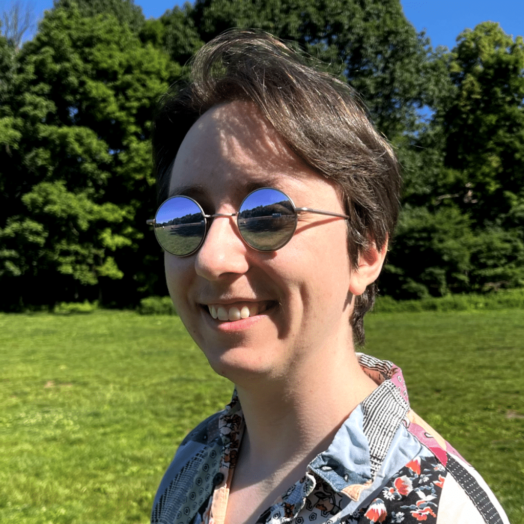 A person smiling outdoors, wearing round sunglasses that reflect a sunny landscape, with green trees in the background.