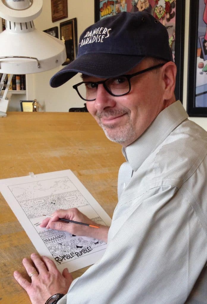 Terry Moore wearing a baseball cap and glasses is sitting at a table, drawing on a piece of comic art with a pencil. The background features framed artwork and a lamp.