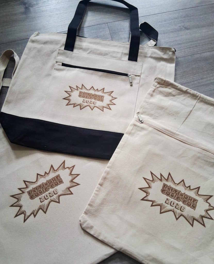 A collection of beige canvas bags featuring the logo for Baltimore Comic-Con 2026, including a large tote with black straps and a smaller bag with a zipper.
