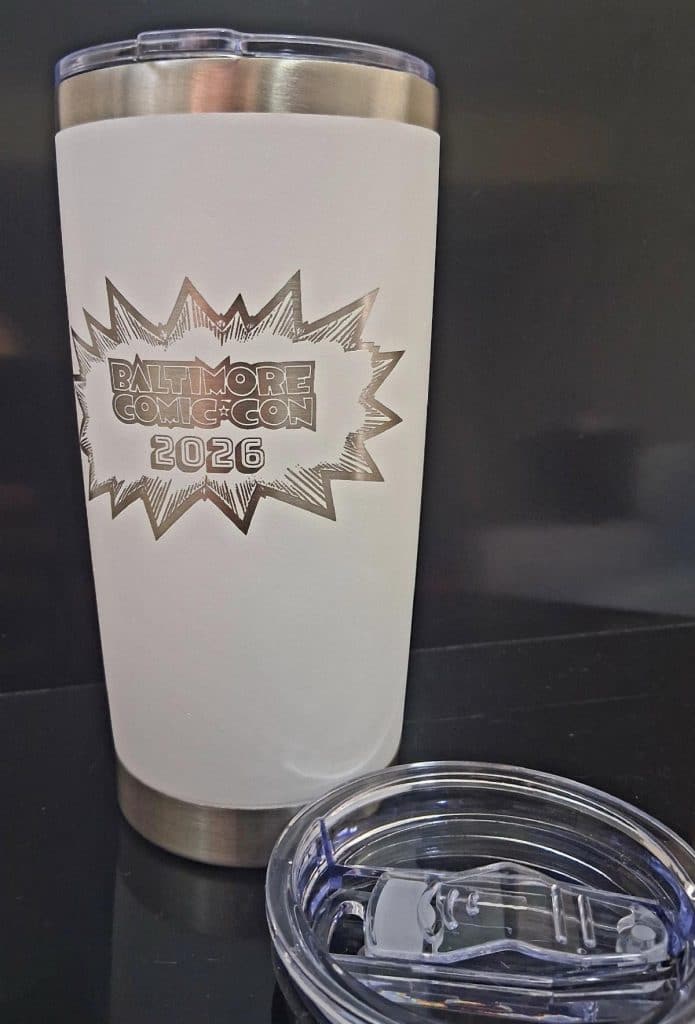A stainless steel tumbler with a white exterior, featuring an engraved design that reads 'Baltimore Comic-Con 2026' inside a starburst graphic, alongside a clear lid with a sliding opening.