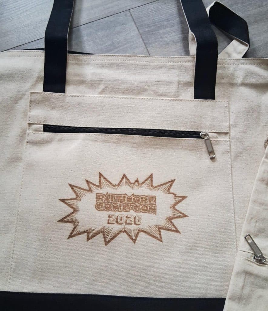 A cream-colored tote bag featuring a zippered pocket and the text 'Baltimore Comic-Con 2026' in a comic-style burst design.