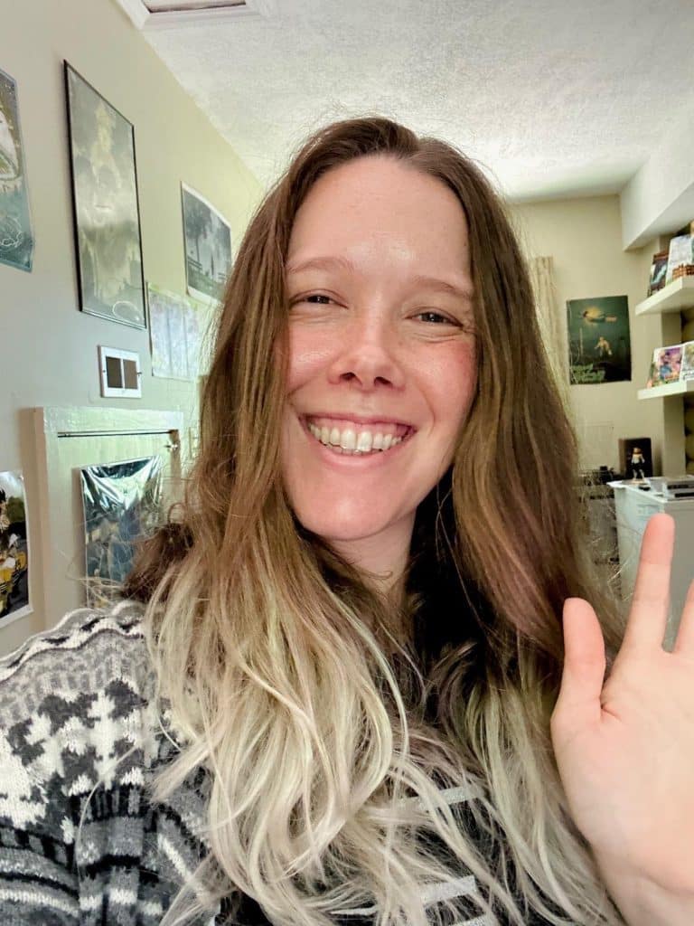 A woman smiling and waving at the camera, with long hair and wearing a patterned sweater, in a well-lit indoor space decorated with various artworks.