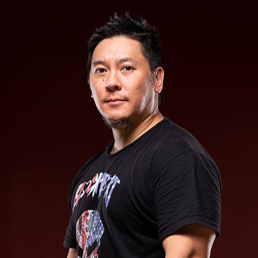 A person standing in front of a dark red background, wearing a black t-shirt with a graphic design, looking directly at the camera.