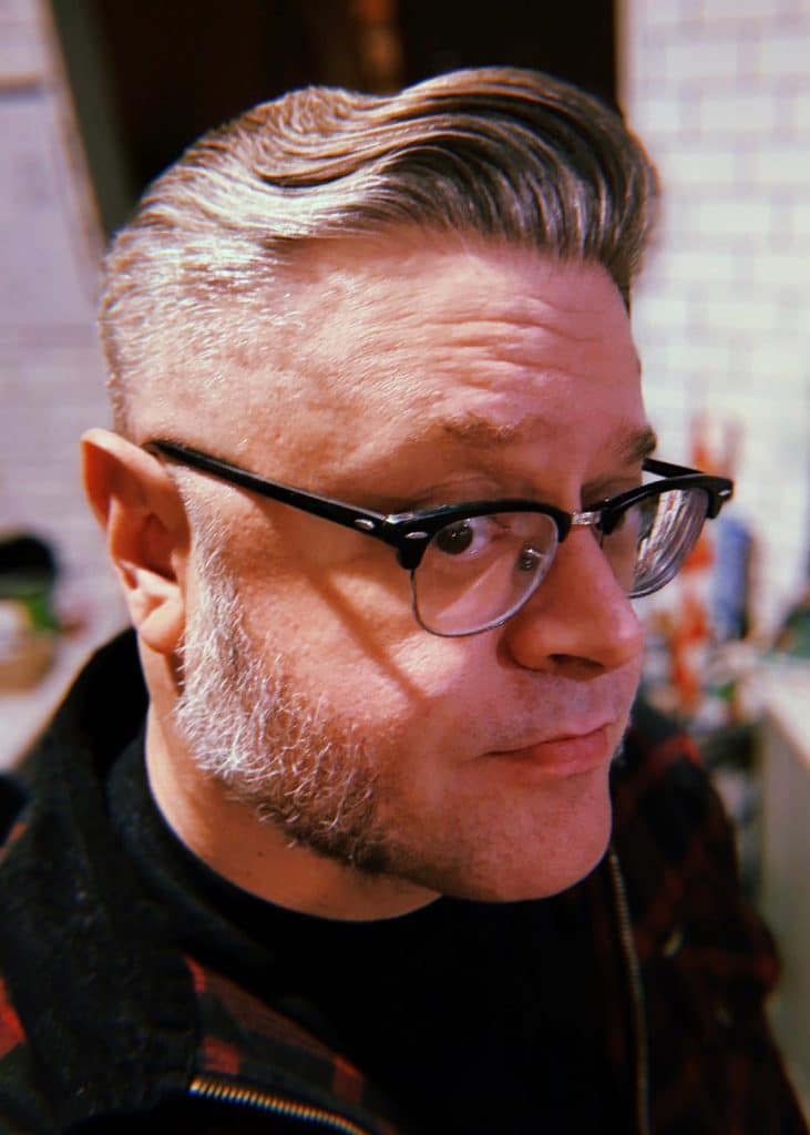 Profile view of a man with slicked-back gray hair and distinctive sideburns, wearing glasses and a black shirt under a plaid jacket.