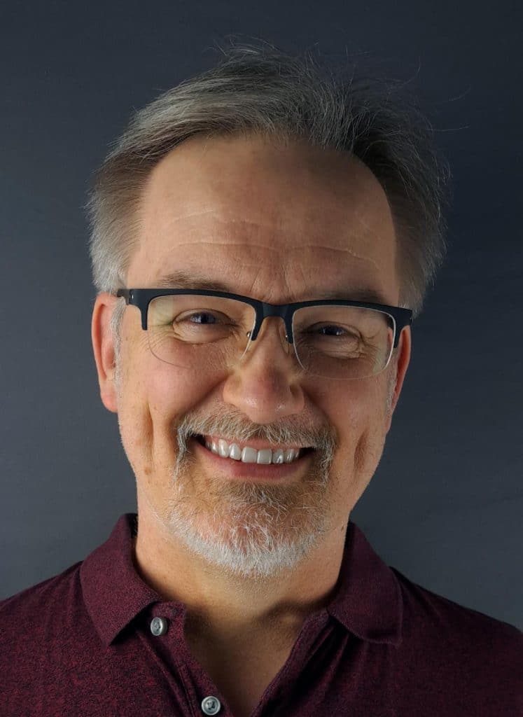 A smiling man with glasses and gray hair, wearing a maroon polo shirt, against a dark background.
