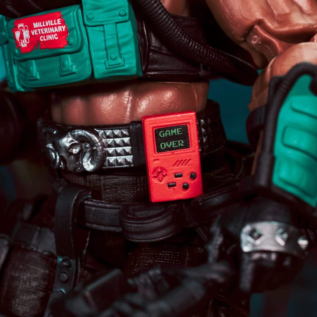 Close-up of a toy figure displaying a red handheld gaming device with 'GAME OVER' on the screen, attached to a textured black belt with studs.