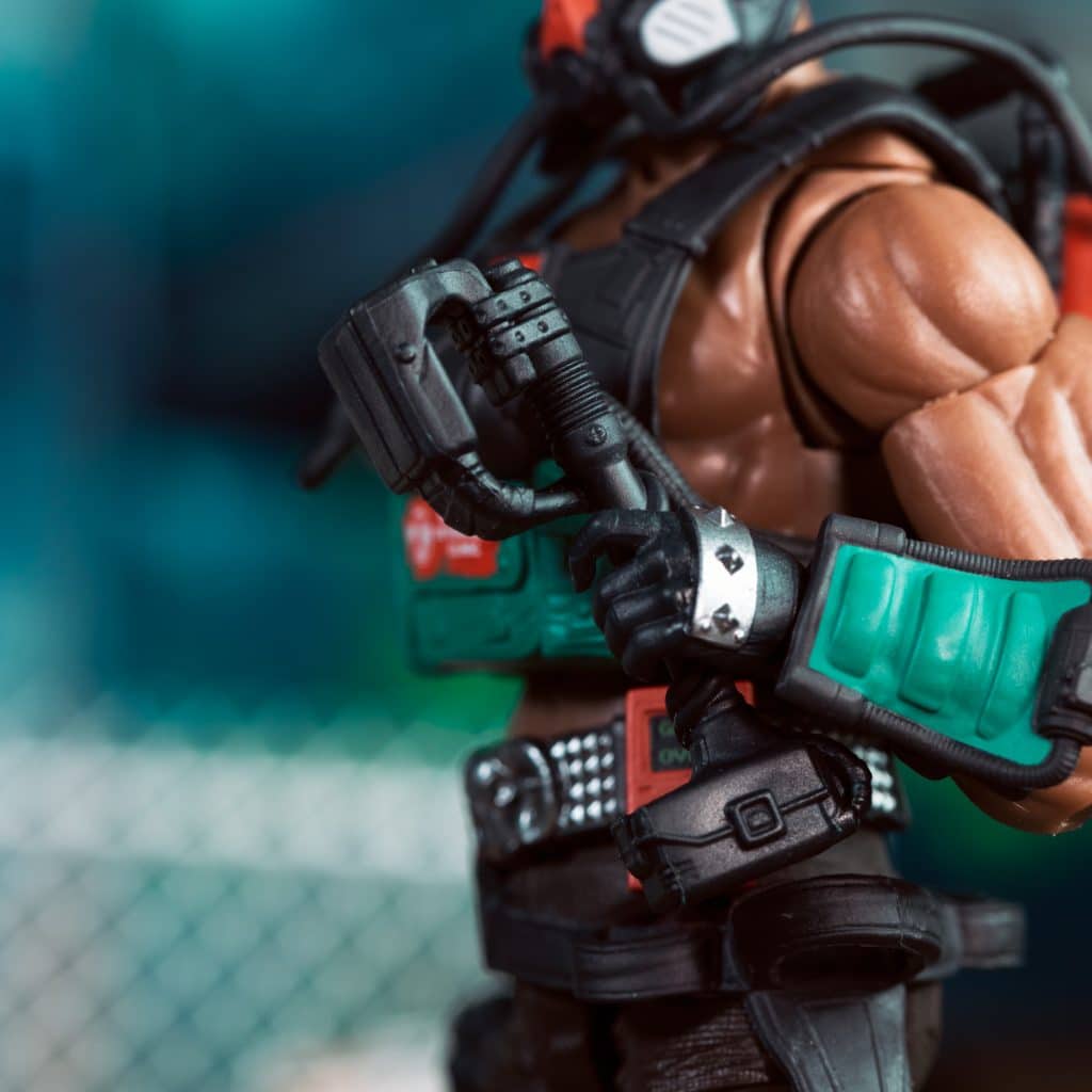 Close-up of a muscular action figure wearing a tactical vest and mask, holding a weapon with cables attached, set against a blurred background.