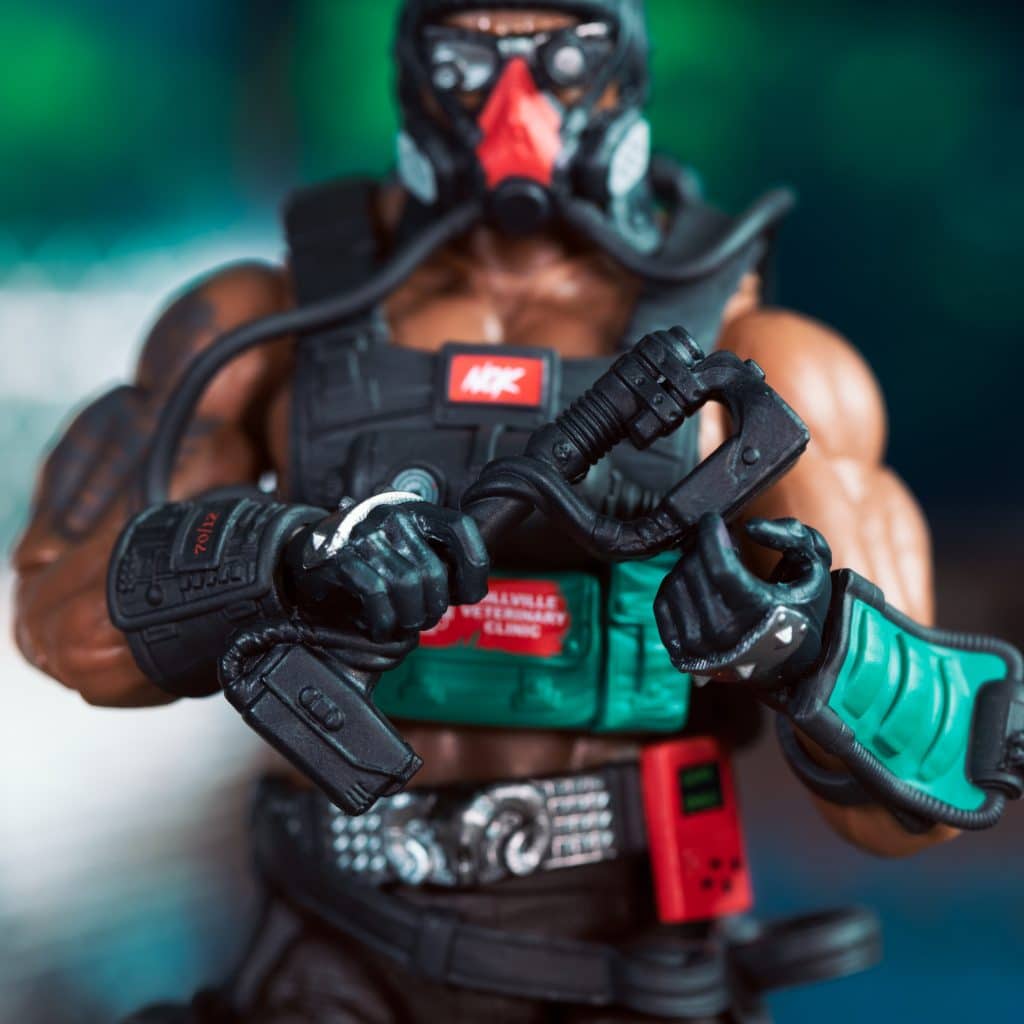 Close-up of an action figure in tactical gear, holding a tool with both hands, set against a blurred background.