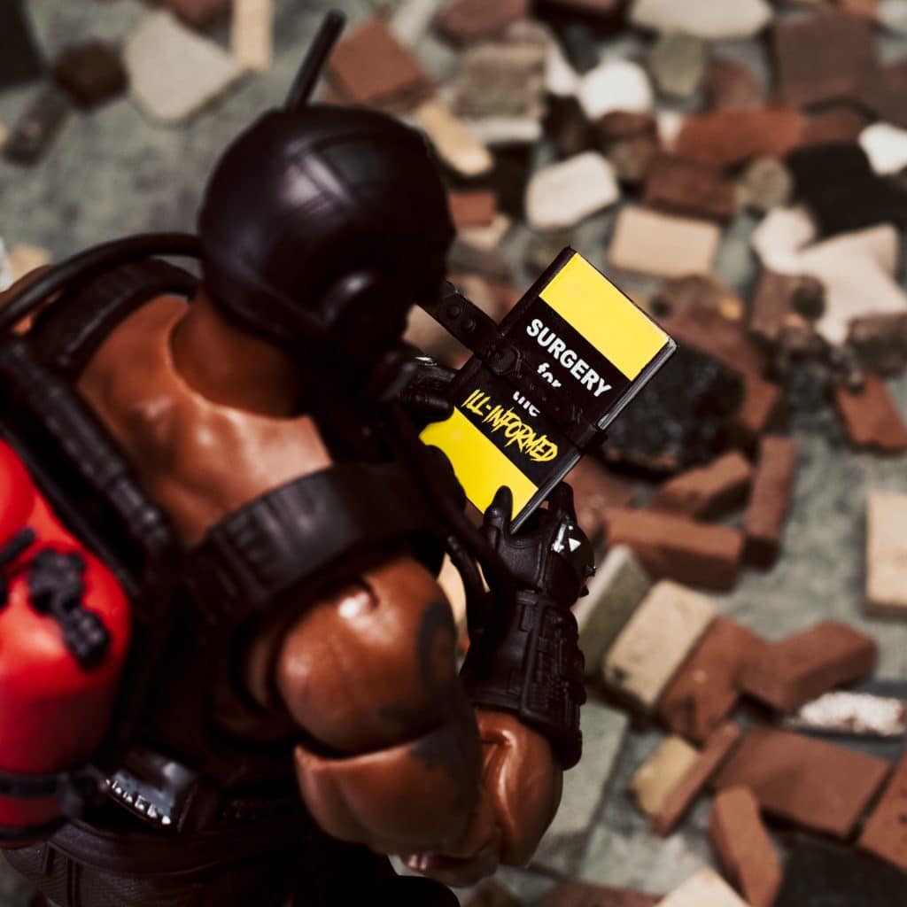 A close-up of an action figure holding a yellow surgical manual titled 'SURGERY' with the subtitle 'ILL-INFORMED', surrounded by a scattered pile of bricks.