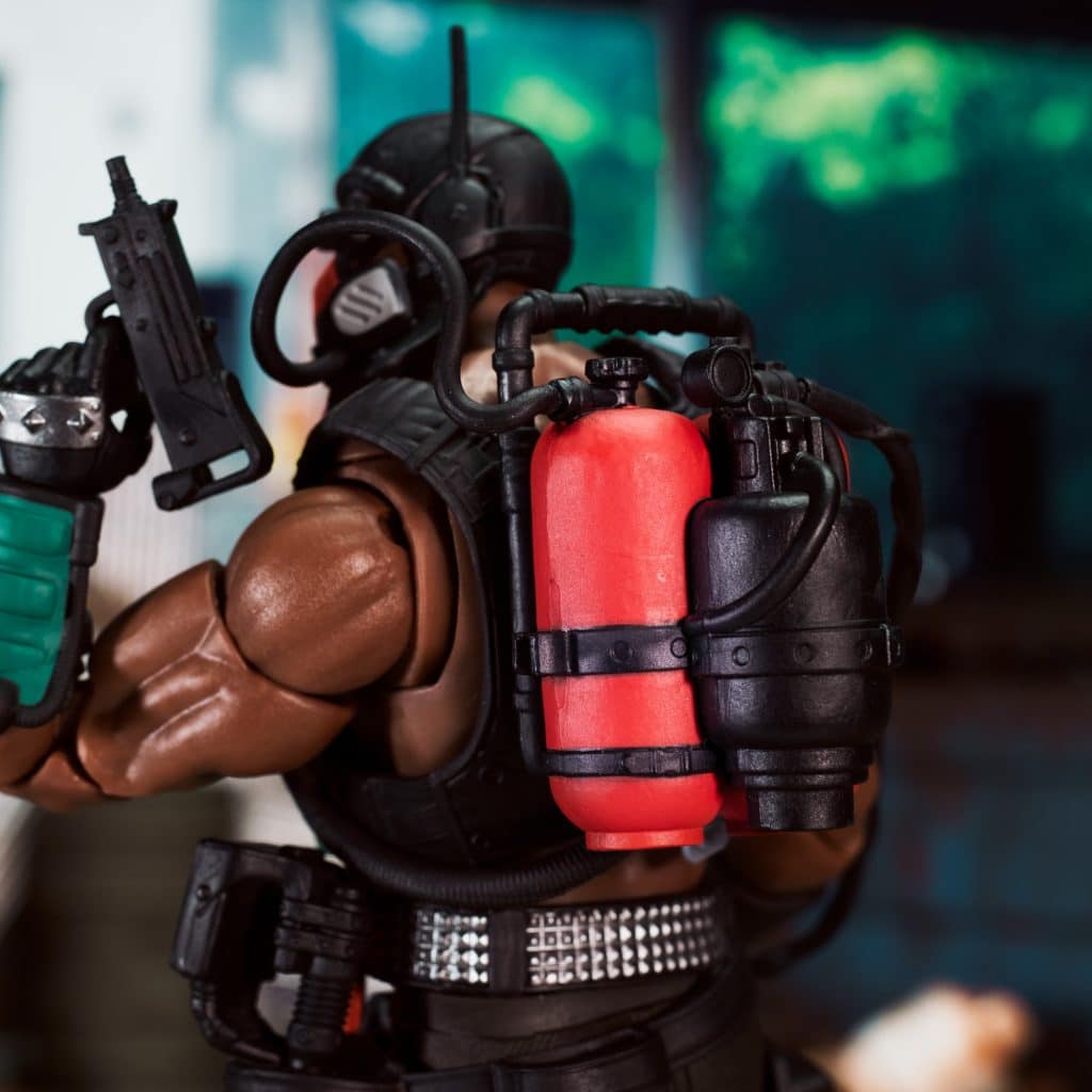 A close-up of an action figure with muscular arms, wearing a tactical outfit, holding a weapon, and featuring a red and black backpack with tanks.
