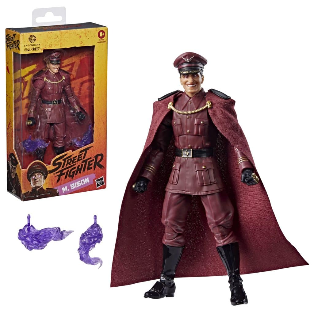 A detailed action figure of M. Bison from Street Fighter, dressed in a military-style uniform and cape, with a packaging box and two purple energy accessory pieces.