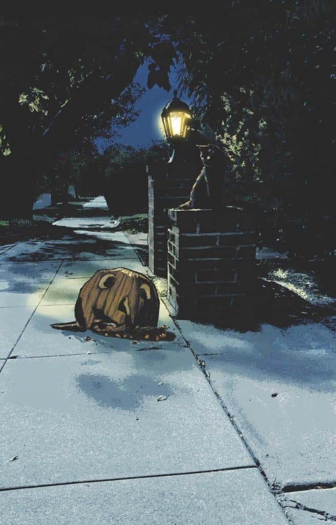 A dimly lit sidewalk at night featuring a street lamp and a cat perched on a brick pillar, with a carved pumpkin laying on the ground.